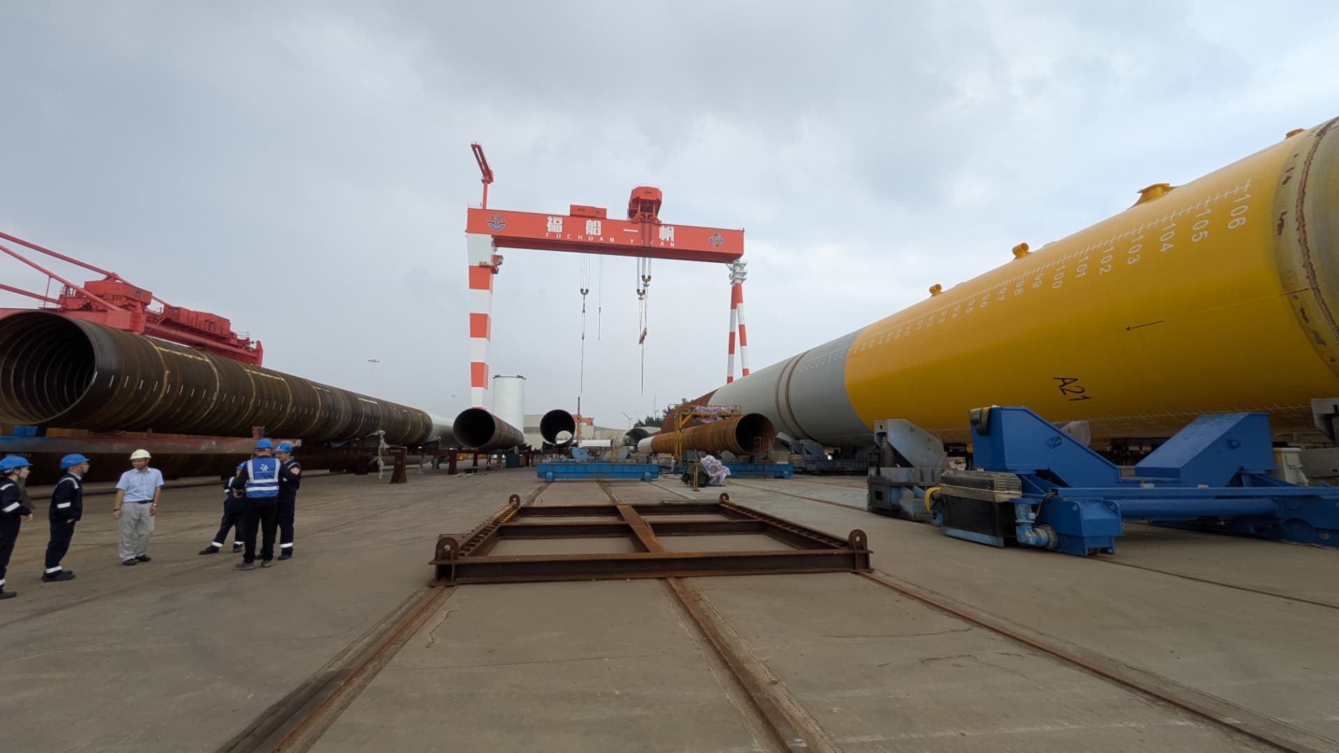 Monopiles and transition pieces staged for load-out under a red and white gantry crane